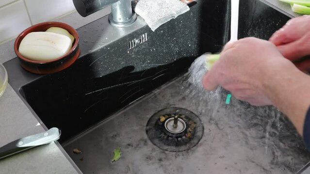 A man cleans celery stalks in a kitchen sink under running water. 