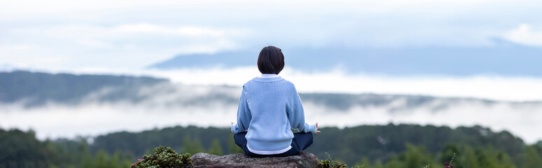 Panorama back view of woman in hoodie is relaxingly practicing meditation yoga at the top of mountain with mist and fog in summer to attain happiness from inner peace wisdom for healthy mind and soul