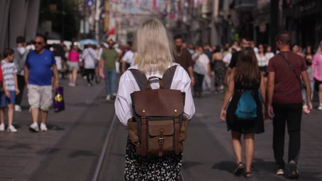 Back view of young beautiful blonde female tourist walking on street, summer fashion style, with backpack and white shirt, travel. Attractive girl exploring new city. Sunny Image Fashion Portrait