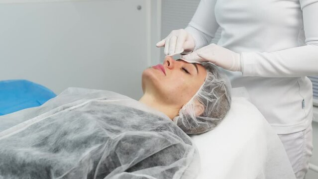 Cosmetologist is applying cream with anesthesia on patient's face skin before biorevitalization procedure. Woman in beauty clinic with doctor beautician preparing to treatment using numbing cream.
