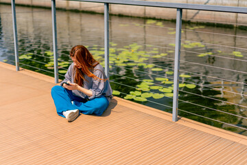 a young red-haired girl sits on a pier or bridge with a phone and communicates with a friend in the messenger of communication in social networks