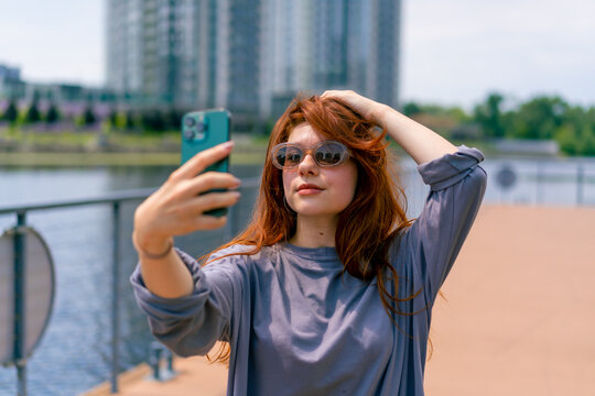 a young red-haired girl in sunglasses with a phone in her hands takes a photo of herself on a selfie camera