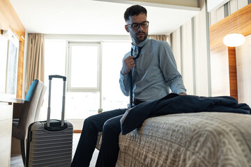 The businessman enters his hotel room, takes a seat onto the hotel bed. The comfort of the room offers a well-deserved moment after a long day of meetings.