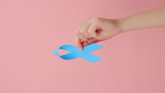 Vertical shot of female hand holding blue ribbon, posing over pink background with copy space for design. Healthcare, International men, Father and World Cancer Day concept. Symbol for support men