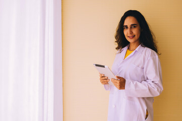 A female doctor stands by the window of the hospital room holding a tablet.