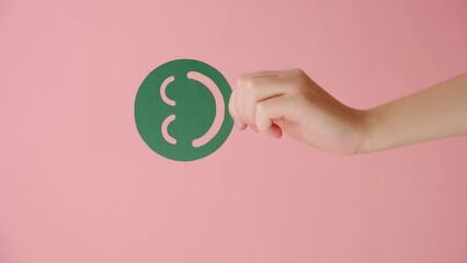 Vertical video of female hand holding positive green smiley face, posing isolated over pastel pink color background wall in studio with copy space for promotion content or design. Emotion concept