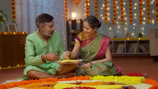 Happy elderly senior couple preparing Diwali flower rangoli for festival celebration at home - concept of traditional custom, relationship bonding and special occasion