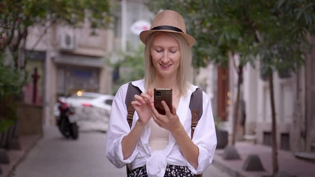 Caucasian attractive female tourist traveler in saloon hat and shirt, looking at smartphone, smiling while walking in city and enjoying vacation. Searches in app for taxis maps with city geolocation