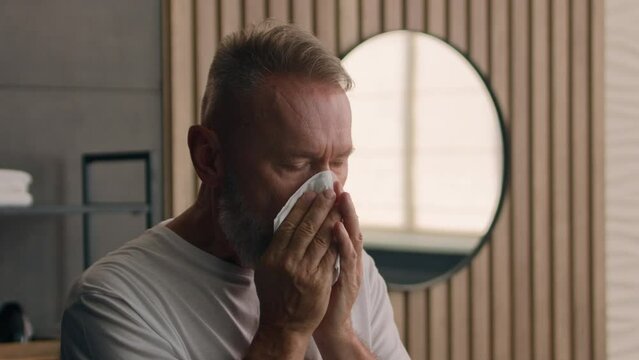 Old Caucasian 60s man sick at home bathroom sneeze wipe nose with paper napkin aged retired male mature senior ill person grandfather sneezing with runny nose cold allergy symptoms respiratory illness