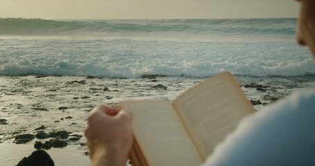Man relaxing on beach and reading a book. Big stormy ocean waves on the background.