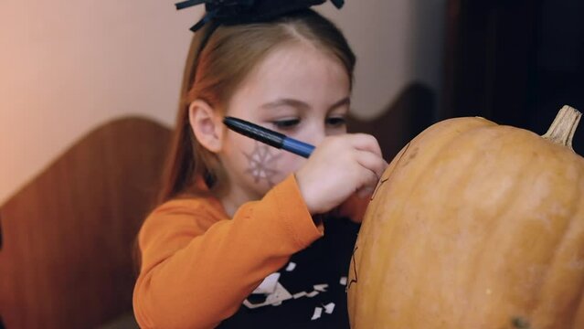 Cute happy little girl preparing for halloween, painting drawing scary face on pumpkin while sitting at table in kitchen at home, smiling child making jack-o-lantern. Holiday decoration concept