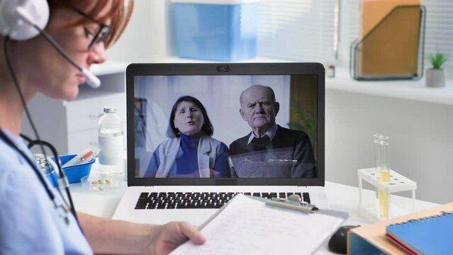 receiving patients online, young female doctor communicates with an elderly man and woman via video conference on a laptop using a headset while sitting in medical office