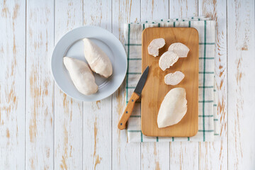 Boiled chicken breast in a plate, slices of meat and knife on cutting board on light wooden table....