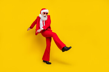 Fototapeta na wymiar Full length photo of cute positive man pensioner dressed red suit santa hat having fun walking empty space isolated yellow color background