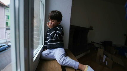 Child nose picking while sitting by window at home looking out from apartment, bored little boy...