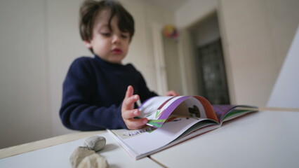 Child turning book pages looking at images and text. Little boy reading kindergarten school material - obrazy, fototapety, plakaty