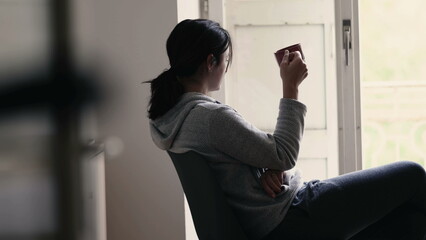 Thoughtful woman sipping warm drink while staring out from home balcony window in contemplation. Person sipping coffee or tea while thinking, starting the day routine