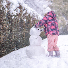 Little girl in warm winter clothes building snowman outdoors in park during snowfall.