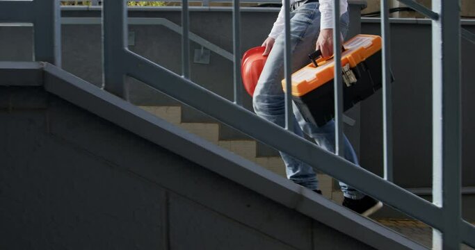 A man in a white shirt climbs the street stairs, carrying a construction worker&rsquo;s helmet and a toolbox in his hands. The engineer climbs the stairs.