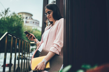 Young woman with papers in hand reading message on phone in city