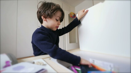 One little boy sitting at desk opening table drawer and taking drawing material, engaged with kindergarten school work at home - obrazy, fototapety, plakaty