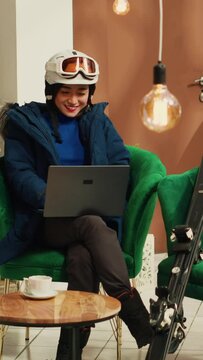 Vertical Video Person reading place reviews on laptop at luxury ski resort, sitting in lobby with skiing gear and accessories. Asian woman working remote on wireless pc before practicing extreme