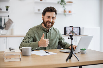 Confident bearded man sitting at desk and working on wireless laptop while recording video on modern smartphone fixed on tripod showing thumb up