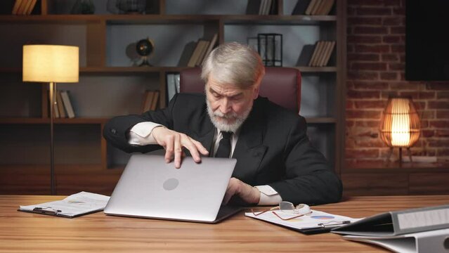 Serious older person in formal suit opening laptop computer after putting of reading glasses in workplace. Confident entrepreneur getting ready for scheduled online meeting in office interior.