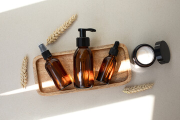 Amber bottles with facial cosmetics on a wooden tray on the beige concrete background with a streak of daylight. Flat lay