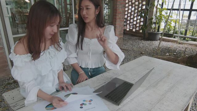 Modern business concept of portrays two pretty young entrepreneurs, in an remote business meeting. Their radiant smiles and cheerful faces with laptop and analysis paper for their success.