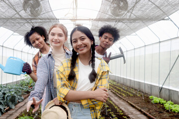 Group of happy Multiethnic teenager friend work in vegetable farm, portrait of smiling young diverse farmer standing together, showing garden tools and watering can in agricultural field greenhouse.