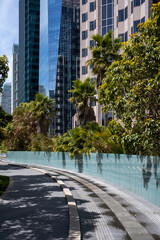 Paved walkway near mirrored fence and exotic plants and palms growing against tall skyscrapers in Salesforce Park located in San Francisco on sunny day