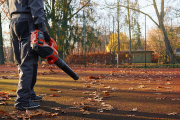 Unrecognizable male worker wearing blue uniform blowing out yellow leaves park area. Cropped view...