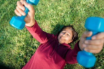 Top view of a senior latin woman doing lying down exercise with dumbbells outdoor on the grass.