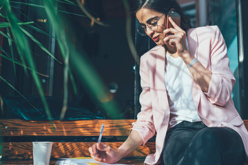 Pensive smiling woman with takeaway coffee noticing in notebook and talking on phone in city