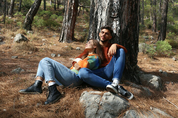 Young couple relaxing under the tree in the woods. Hipster boyfriend and girlfriend leaning on the tree trunk and hugging. Close up, copy space, forest background.