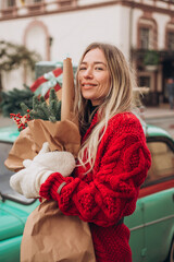 Young beautiful blonde woman in a red knitted cardigan and warm mittens holds a Christmas bouquet while walking through an old European city