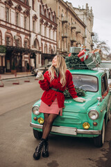 Young blonde woman holding a glass of champagne while sitting on the hood of a retro car decorated with Christmas boxes