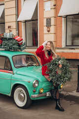 Beautiful young blonde woman in a red knitted cardigan holding a Christmas wreath while sitting on the hood of a retro car