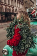 A blonde girl in a red knitted cardigan carries a Christmas wreath on her shoulder