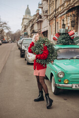 A blonde girl in a red knitted cardigan carries a Christmas wreath on her shoulder