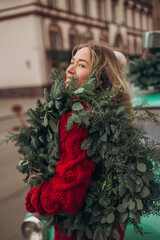 A blonde girl in a red knitted cardigan carries a Christmas wreath on her shoulder