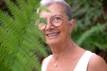 Portrait of happy cheerful senior woman in trekking day in the forest partially hidden by a fern...