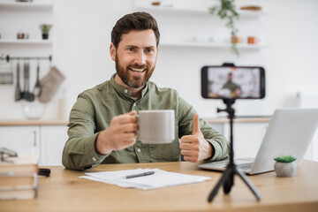 Happy attractive streamer sitting at table near modern laptop and recording video content on...