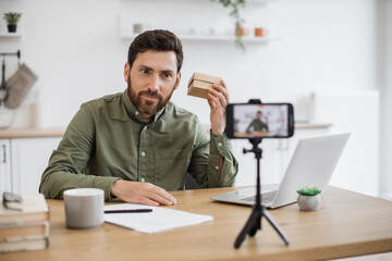 Caucasian bearded man using smartphone and tripod for live broadcasting at home. Friendly male influencer sitting at desk and holding small parcel box near ear.