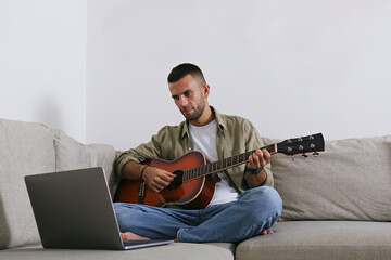 Bearded guy taking an online lesson on playing the acoustic guitar guitar on his laptop. Close up,...