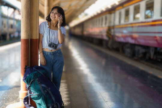 An Asian female tourist is using a mobile phone while wearing a film camera around her neck and a backpack on the platform of a train station.