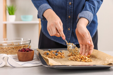 Making granola bars. Woman putting mixture of oat flakes, dry fruits and other ingredients onto baking tray at table in kitchen, closeup