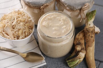 Spicy horseradish sauce in jars, roots and spoon on grey wooden table, closeup