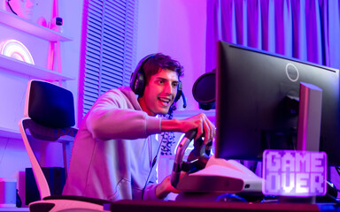 Handsome happy young male game online streamer, caster smiling with fun, succeed for winner, using wheel to play, compete driving game, sitting in room with decorated neon light. Looking at computer.